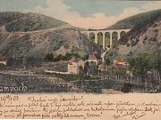 Reprodukce pohlednice Žampachu z počátků provozu tratě. Snímek zachycuje dokončený most ze strany od řeky Sázavy. Po mostě právě projíždí vtipně dokreslený vlak z Jílového směrem do Vraného nad Vltavou.
